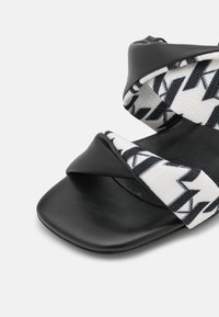 Sandales en cuir noir avec design croisé, dotées d'une sangle en tissu tissé au motif géométrique blanc et noir. Bout carré et semelle plate.