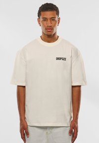 Cremefarbene Baumwoll-T-Shirt mit kurzen Ärmeln und rundem Ausschnitt. Mit einem schwarzen "DROPSIZE" Textlogo auf der Brust.