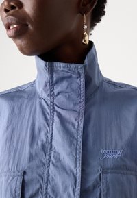 Svijetloplena košulja s kragnom koja ima dvije prednje džepove i izvezeni logo "tommy jeans", izrađena od teksturirane tkanine s vidljivim detaljima šavova.