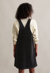 Robe en velours côtelé noir avec des bretelles réglables, de forme A-line et d'une texture douce. Portée par-dessus une chemise à carreaux légère à manches longues.
