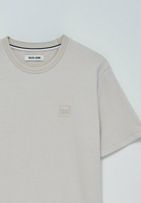 Camiseta corta de manga con textura en color beige claro, con cuello redondo y pequeño parche cuadrado con el logo "Salsa Jeans" en el lado izquierdo del pecho.