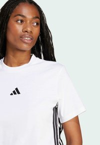 Valkoinen lyhythihainen t-paita, jossa on musta Adidas-logo rinnassa ja kolme mustaa raitaa sivuilla. Sileä puuvillakangas.