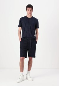 Camiseta negra de manga corta con estampado, combinada con pantalones cortos oscuros que tienen una cintura elástica y cordón. El modelo lleva zapatillas blancas.