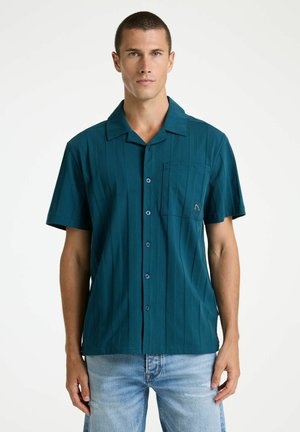 Moški s kratkimi lasmi, oblečen v turkizno kratko srajco z gumbi in svetlo modre kavbojke, stoji pred belim enobarvnim ozadjem.