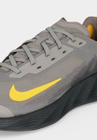 Γκρι αθλητικό παπούτσι με κίτρινο λογότυπο Nike, γκρι κορδόνια και υφή μαύρης σόλας, σε γωνία που δείχνει πλευρικές και επάνω λεπτομέρειες.