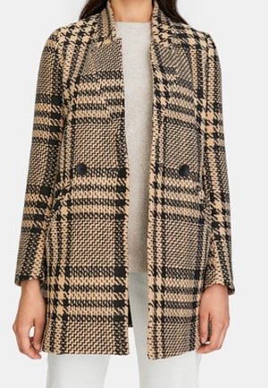 Beige and black plaid double-breasted coat with black buttons worn over a light gray top and white pants.