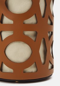 Brown leather holder with a geometric cutout design, revealing a light beige textured inner fabric. Circular and angular shapes are prominent.
