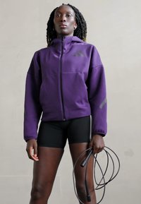 Casaco roxo com fecho éclair e capuz, com detalhes cinzentos, combinado com calções pretos. O modelo segura uma corda de saltar, exibindo um visual desportivo.
