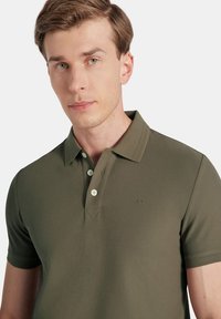 Oliwkowa koszulka polo z teksturowanej tkaniny, dwoma guzikami przy kołnierzu i subtelnym logo po lewej stronie klatki piersiowej. Krótkie rękawy.