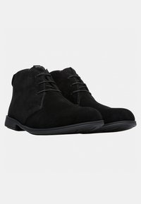 Un paio di stivali neri in suede fino alla caviglia con lacci, punte arrotondate e suole in gomma piatta su uno sfondo bianco.