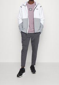Weiße und graue Zip-Jacke mit Kapuze, rosa T-Shirt, graue schmal geschnittene Hose und schwarze Sportschuhe. Mit Logo und Farbabstufungsdesign.