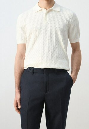 Mann, der ein weißes kurzärmeliges Strick-Poloshirt in schwarze, maßgeschneiderte Hose gesteckt trägt, mit einer Hand in der Tasche.
