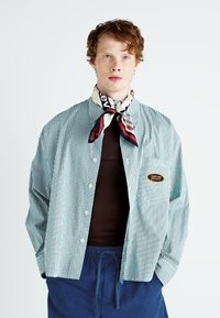 Stripete skjorte i lyseblå og hvit med løs passform, knappestolpe foran, og en lapp på brystet; kombinert med et mønstret skjerf.