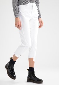 Valkoiset lyhyet farkut, joissa on repaleiset lahkeet, yhdistettyinä kiiltävän mustiin nilkkabootseihin. Asusteena on mustavalkoruudullinen pitkähihais shirt.