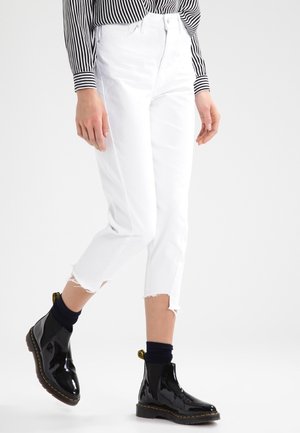 Jean blancs raccourcis avec ourlet effiloché, associés à des bottines noires brillantes. La tenue comprend une chemise à rayures noires et blanches à manches longues.