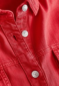 Zbliżenie na czerwoną tkaninę dżinsową z metalowymi guzikami oznaczonymi "Next Jeans Since 1982" oraz widocznym ściegiem dziurki na guzik.