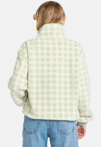 Person viewed from behind wearing a light green checked fleece jacket and blue jeans, with long hair and sunglasses.