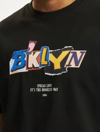 Black t-shirt featuring a colorful graphic design with cut-out letters spelling "BKLYN" and a slogan beneath in white text.