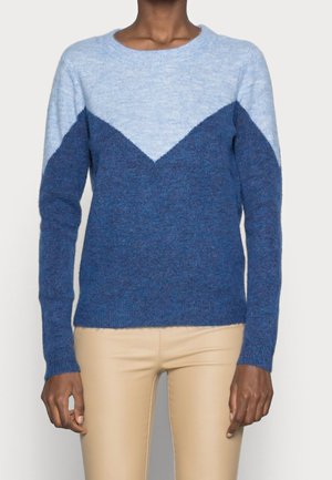 Kobieta ubrana w sweter z długim rękawem, z jasnoniebieską górną częścią i wzorem jodełki w ciemnoniebieskim kolorze, w połączeniu z beżowymi spodniami.