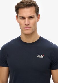 Camiseta de algodón de manga corta en azul marino, con un logo blanco y rojo en el lado izquierdo del pecho. Cuello redondo con una textura suave. Diseño liso.