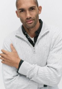 Ljust grått stickat tröja med dragkedja i halsen, med ett texturerat kabelmönster och ribbade ärmslut i mörkgrön.