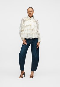 Γυναίκα με πλεξούδες, ντυμένη με διάφανη λευκή μπλούζα με βολάν, σκούρο μπλε τζιν και μαύρα πέδιλα με λουράκια και ψηλό τακούνι, στέκεται μπροστά σε λευκό φόντο.