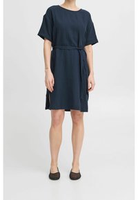 Mörkblå, kortärmad klänning med bälte, tillverkad av strukturerat material; har sidoöppningar och en avslappnad passform. Bärs med svarta, platta skor.