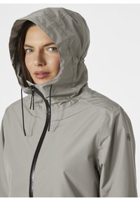 Chaqueta impermeable de color gris claro con capucha ajustada, cremallera negra y cordones ajustables. Textura suave con diseño minimalista.