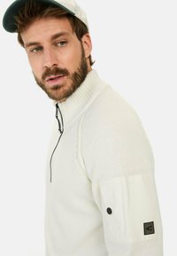 Weißer Reißverschluss-Pullover mit geripptem Kragen und strukturiertem Strick. Mit einem schwarzen Akzent-Patch am Ärmel und einem schwarzen Reißverschluss.