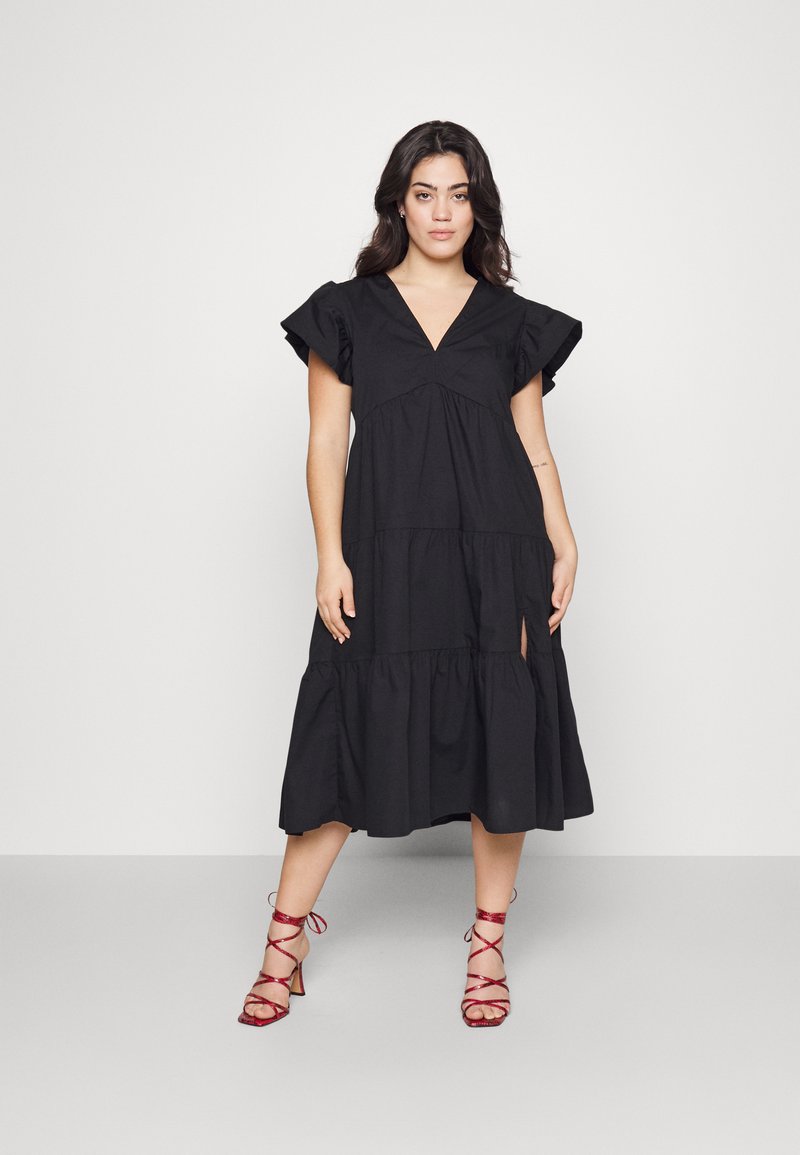 Black cotton dress with a V-neck, short ruffled sleeves, tiered hem, and side slit; worn with red strappy high-heeled sandals.