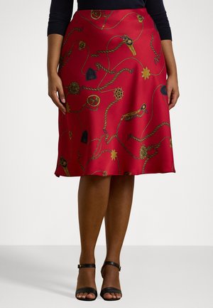 Femme portant une jupe rouge en satin longueur genou avec un imprimé chaîne dorée, noire et nautique, associée à des escarpins noirs à bride cheville.