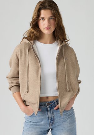 Jeune femme aux cheveux bruns ondulés portant un sweat à capuche beige zippé par-dessus un t-shirt court blanc et un jean bleu clair, mains dans les poches.
