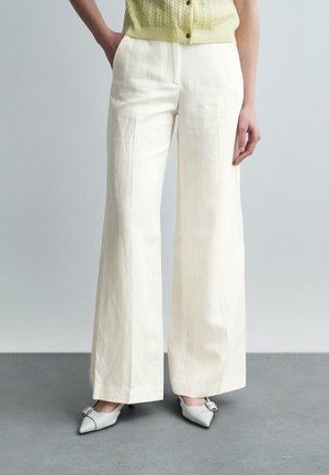 Žena oblečená vo vysokých, širokých krémovo sfarbených nohaviciach s jemnými vertikálnymi pruhmi, doplnených bielymi lodičkami so zúženou špičkou a prackami.