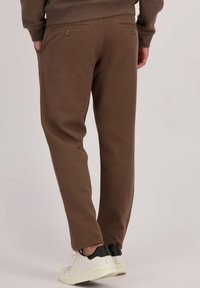 Man in een paarse achtergrond, gekleed in bruin op maat gemaakte broeken en witte sneakers, met zijn handen in zijn zakken en met zijn rug naar de camera.