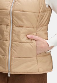 Beige puffer vest met horizontale stiksels, volledige voozip, zijzakken en een zachte textuur, gedragen over een witte trui.