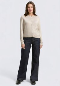 Lichtbeige vest met lange mouwen, ronde hals, knoopsluiting aan de voorkant, gecombineerd met donkerblauwe jeans met wijde pijpen en zwarte schoenen. Eenvoudig ontwerp.