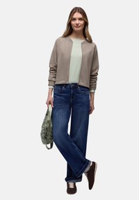 Veste bomber beige clair portée sur un haut vert menthe, associée à un jean bleu ample et des baskets marron foncé. Sac gris en bandoulière.