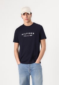 Tamno plava pamučna majica s bijelim logom "HILFIGER USA 1985". Uparena s svijetloplavim trapericama i bež bejzbol kapom. Kratki rukavi.