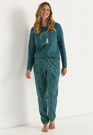 Smilende kvinde står barfodet iført en teal langærmet top med katte- og månemotiv og matchende bukser med blomsterprint.
