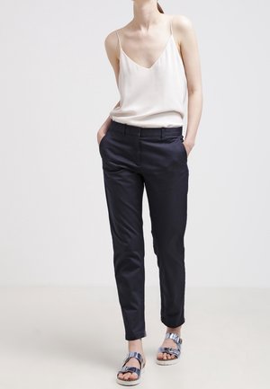 Woman wearing sleeveless cream blouse, black tailored pants, and silver sandals standing against a plain light background.