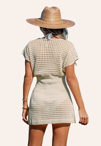 Abito da spiaggia in maglia beige con design a rete aperta, vita aderente e orlo fluente. Abbinato a un cappello di paglia a tesa larga.