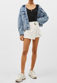 Denim jacket, ribbed black tank top, white wrap shorts with cargo pockets and a silver circle fastener, paired with chunky white sneakers.