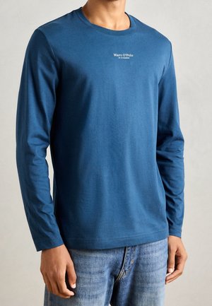 Homme portant une chemise bleue à manches longues avec le texte "Marc O’Polo" sur la poitrine et un jean en denim bleu clair sur fond uni.