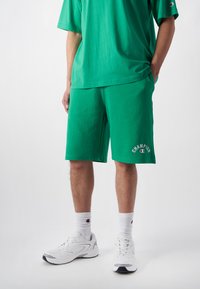 Grüne Baumwollshorts mit einem elastischen Bund, versehen mit einem weißen "Champion"-Logo. Kombiniert mit einem passenden grünen T-Shirt und weißen Sneakern.