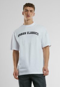 Balta medvilninė marškinėlių palaidinė su „URBAN CLASSICS“ užrašu ryškiu juodu tekstu per krūtinę, trumpomis rankovėmis ir laisvu siluetu.