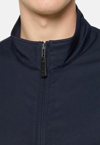 Temno modra zip-up jakna z visokim ovratnikom, izdelana iz gladkega blaga. Ima črno zadrgo z blagovno znamko "SIGNAL" na držalu.