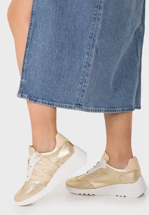 Jambes portant une jupe en denim bleu et des baskets basses dorées avec des lacets blancs et des semelles épaisses blanches striées sur fond blanc.