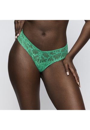 Kobieta ubrana w zieloną koronkową bieliznę w kwiatowe wzory, ręka spoczywa na biodrze, pokazując środkową część ciała i górne części ud na neutralnym tle.