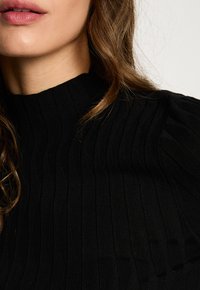 Unteres Gesicht einer Frau mit natürlichen Lippen, welligem braunem Haar und schwarzem, geripptem Rollkragenpullover vor einem neutralen Hintergrund.