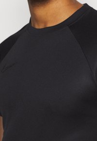 Camiseta deportiva negra con cuello redondo y mangas cortas de tipo raglán. Textura suave con un sutil detalle de logo en el pecho.